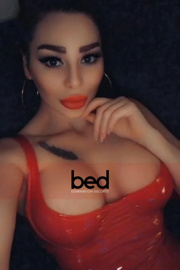 Mistress Allya taking a selfie wearing a red PVC bra