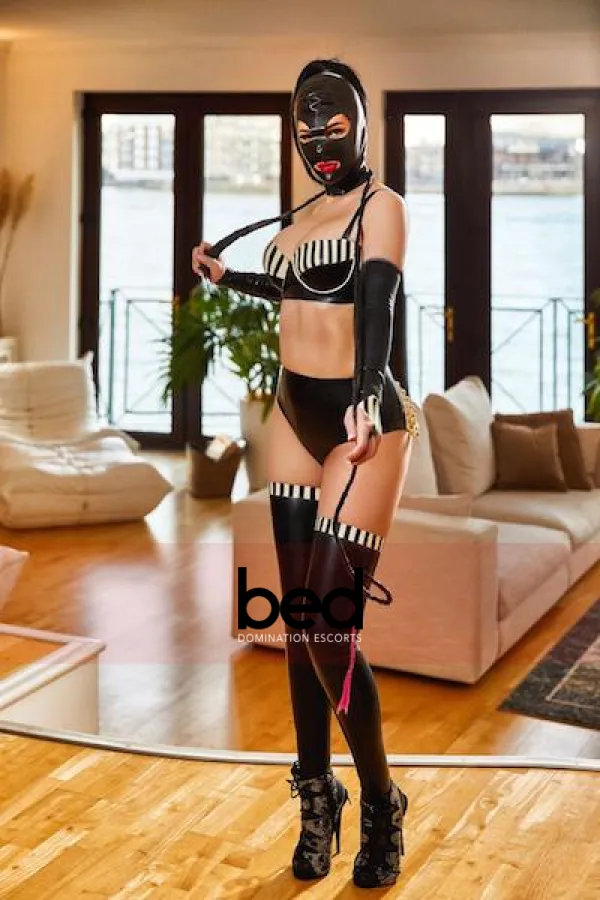 Jade standing in her leather mask and lingerie as she is holding a whip 